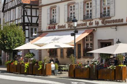 The terrace of a traditional hotel-restaurant, with parasols and flower-filled planters, situated on a picturesque street lined with half-timbered buildings