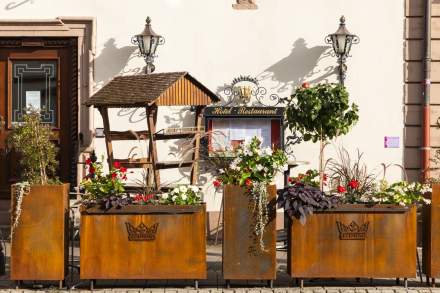 Devanture d’un hôtel-restaurant avec deux lampadaires muraux, un présentoir en bois pour menus et plusieurs jardinières en acier rouillé contenant des plantes et fleurs colorées