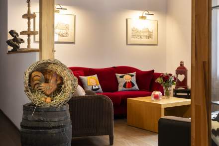 Salon chaleureux avec canapé rouge décoré de coussins illustrés, table basse en bois avec bougie et bouquet de fleurs, deux cadres accrochés au mur, et en premier plan une décoration rustique avec un coq en paille posé sur un tonne