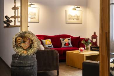 Salon chaleureux avec canapé rouge décoré de coussins illustrés, table basse en bois avec bougie et bouquet de fleurs, deux cadres accrochés au mur, et en premier plan une décoration rustique avec un coq en paille posé sur un tonne