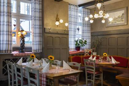Salle de restaurant traditionnelle avec tables en bois, nappes à carreaux rouges, chaises grises, serviettes pliées, bouquets de tournesols, grandes fenêtres avec rideaux à carreaux et lustres décoratifs
