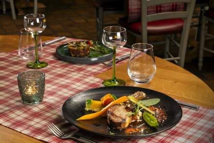Assiette gastronomique avec viande rôtie, légumes colorés et sauce, présentée sur une table dressée avec verres à pied et bougie décorative dans un restaurant.