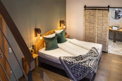 Chambre moderne avec lit double, tête de lit en bois, coussins verts, plaid à motifs, éclairage mural chaleureux et porte coulissante en bois menant à une salle de bain avec carrelage décoratif.