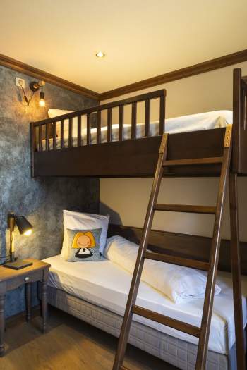 Chambre avec lits superposés en bois sombre, lit inférieur avec coussin décoratif illustré d'un personnage, lampe de chevet noire sur une table en bois, mur texturé gris et éclairage chaleureux.