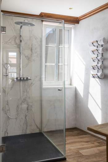 Cabine de douche moderne avec paroi en verre, mur en marbre clair, pommeau de douche pluie, fenêtre apportant lumière naturelle et porte-serviettes mural avec serviettes roulées.