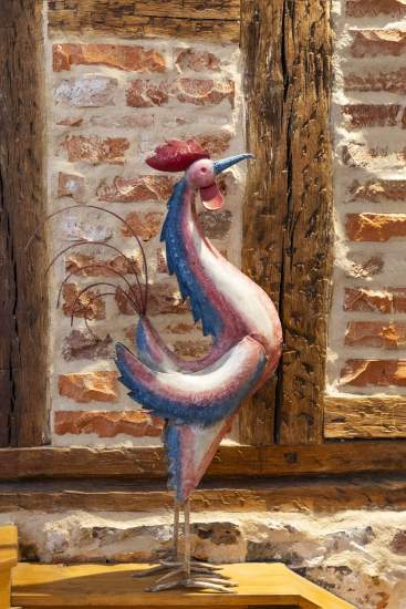 Sculpture décorative en forme de coq coloré, avec des teintes rouges, bleues et blanches, posée devant un mur en briques et poutres en bois rustiques.