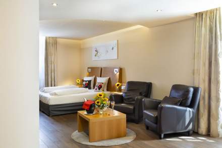 Chambre avec deux lits simples côte à côte, draps blancs, coussins décoratifs à motifs rouges, mur en pierre apparente, deux lampes murales noires et fenêtre avec rideaux gris.