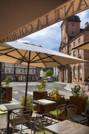 Terrasse d’un café avec parasols donnant sur une place circulaire, entourée de bâtiments historiques en pierre et maisons à colombages, sous un ciel partiellement nuageux.