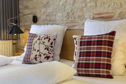 Chambre avec deux lits simples côte à côte, tête de lit en bois, mur en pierre apparente, coussins rouges à motifs, et fenêtre avec rideaux gris