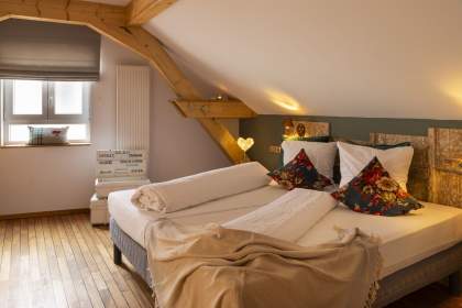 Chambre sous combles avec lit double, deux oreillers décorés de housses à motifs floraux, couverture beige, poutres en bois apparentes, fenêtre avec store gris et sol en parquet